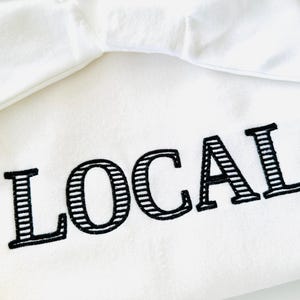 May include: White hooded sweatshirt with the word "LOCAL" embroidered in black with a striped pattern.