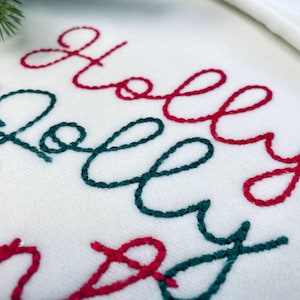 Puede incluir: Primer plano de una tela blanca con texto bordado en rojo y verde. El texto dice "Holly Jolly Season" en una fuente cursiva, con una ramita de vegetación en la esquina superior izquierda. Decoración festiva navideña.