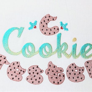 May include: Embroidered design of the words "Cookie Tester" with a cookie-shaped letter "C" and holly sprigs. The letters "Tester" are made of cookie shapes with bite marks.