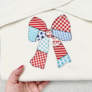 May include: A white fabric item with a large, embroidered bow. The bow features a patchwork design in red, white, and blue, with patterns including stars, stripes, and checks. The item is held by a hand with red nail polish.