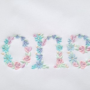May include: Embroidered floral design of the word "one" in pastel pink, blue, and green colours. The design is perfect for a baby's first birthday.