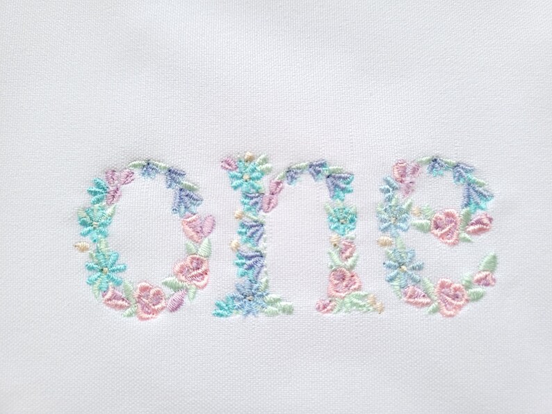 May include: Embroidered floral design of the word "one" in pastel pink, blue, and green colours. The design is perfect for a baby's first birthday.