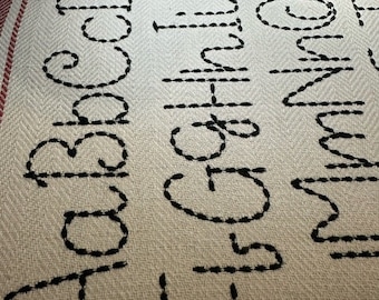May include: A white fabric with black embroidered letters of the alphabet. The letters are stitched in a simple outline style.