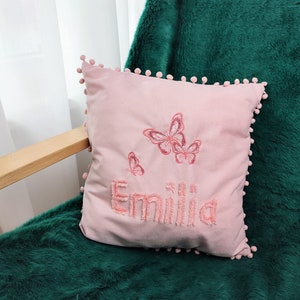 K&ouml;nnte beinhalten: Ein rosa Kissen mit Pompon-Besatz und gestickten Schmetterlingen sowie dem Namen "Emilia".