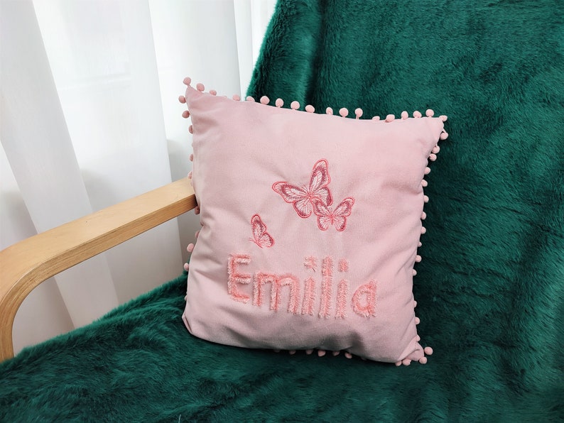 K&ouml;nnte beinhalten: Ein rosa Kissen mit Pompon-Besatz und gestickten Schmetterlingen sowie dem Namen "Emilia".