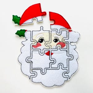 May include: A felt Christmas puzzle in the shape of Santa Claus's face. The puzzle is white with black outlines and features a red hat, rosy cheeks, and a white beard. The puzzle is partially assembled.