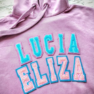 Puede incluir: Una sudadera con capucha rosa con el nombre "LUCIA ELIZA" bordado en letras azules y rosas.