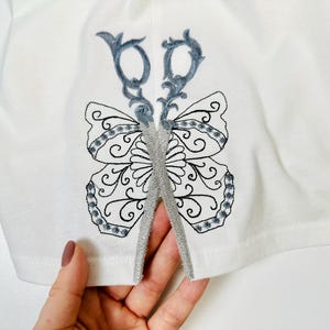 Op de afbeelding: Wit T-shirt met een zilveren geborduurd vlinderontwerp. De vlinder heeft een schaar in het midden.