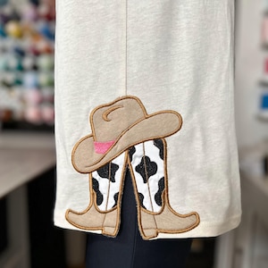 May include: A white shirt with a brown cowboy hat and boots with a black and white cow print design embroidered on the front.