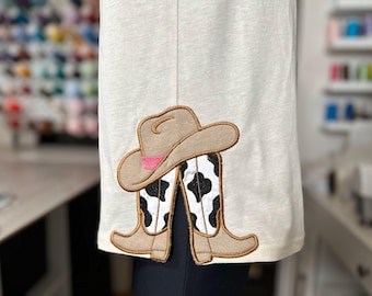 Könnte beinhalten: Ein weißes Hemd mit einem braunen Cowboyhut und Stiefeln mit einem schwarz-weißen Kuhmuster, das auf der Vorderseite gestickt ist.