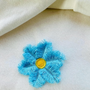 May include: A blue flower with a yellow center, made of fabric with a fringed edge. The flower is on a white background.