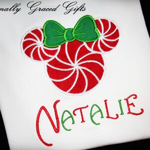 May include: White t-shirt with a red and white candy cane design and a green bow. The name "NATALIE" is embroidered in red and green.
