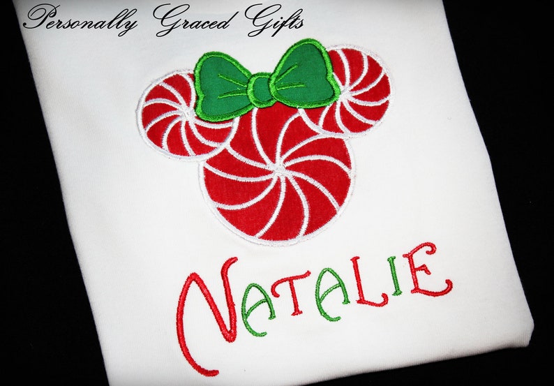 May include: White t-shirt with a red and white candy cane design and a green bow. The name "NATALIE" is embroidered in red and green.