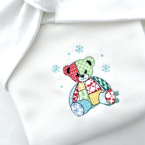 May include: White baby blanket with an embroidered patchwork teddy bear design. The bear features red, green, yellow, and blue patches, with snowflakes scattered around it. The blanket has a folded edge.