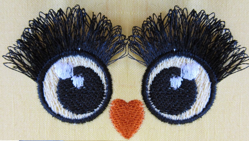 May include: Two black embroidered eyes with white highlights and long black eyelashes, and a red heart embroidered on a yellow fabric.