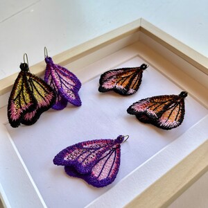 May include: Three pairs of butterfly-shaped earrings displayed in a wooden frame. The earrings are made of delicate lace and feature a variety of colors, including purple, pink, orange, and black.