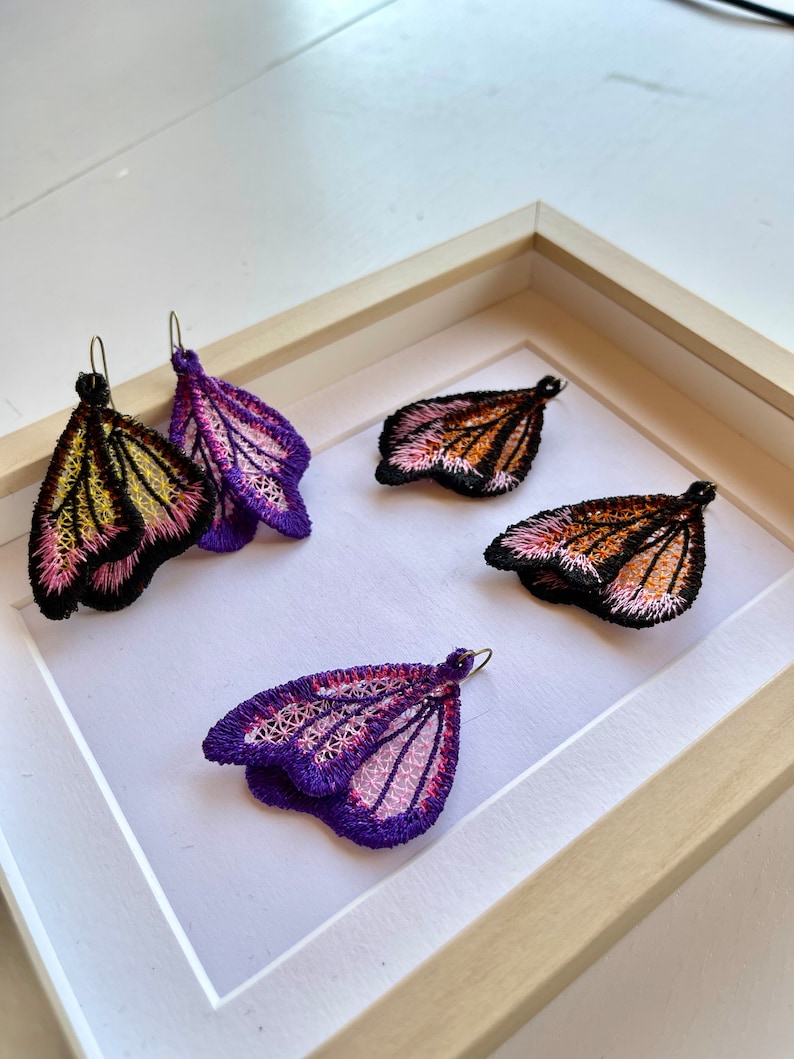 May include: Three pairs of butterfly-shaped earrings displayed in a wooden frame. The earrings are made of delicate lace and feature a variety of colors, including purple, pink, orange, and black.
