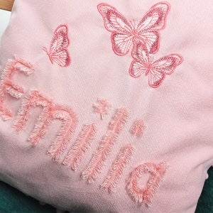 K&ouml;nnte beinhalten: Ein rosa Kissen mit dem Namen "Emilia", der in rosa mit einer flauschigen Textur gestickt ist. Das Kissen ist mit drei rosa gestickten Schmetterlingen verziert.