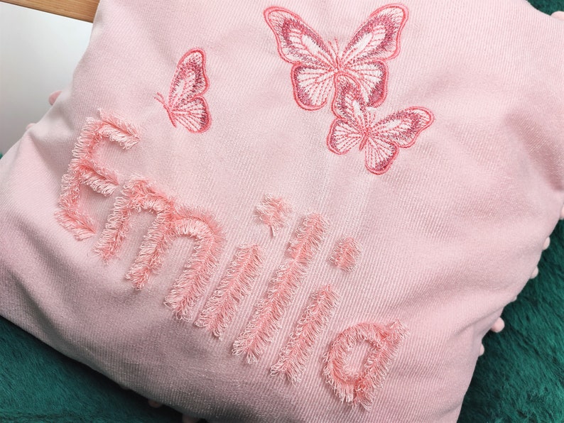 K&ouml;nnte beinhalten: Ein rosa Kissen mit dem Namen "Emilia", der in rosa mit einer flauschigen Textur gestickt ist. Das Kissen ist mit drei rosa gestickten Schmetterlingen verziert.