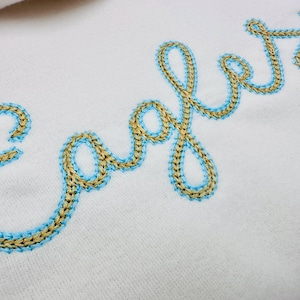 Puede incluir: Primer plano de tela blanca con la palabra "Eagles" bordada en estilo cursivo. El bordado presenta un hilo de color dorado delineado con un hilo azul claro, creando un efecto decorativo y texturizado.