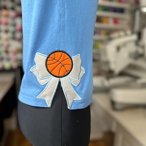 Könnte beinhalten: Ein hellblaues T-Shirt mit einer weißen Schleife und einem orangefarbenen Basketball in der Mitte. Die Schleife ist mit weißem Garn bestickt.