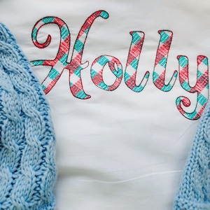 May include: A white shirt with the name "Holly" embroidered in red and teal plaid.