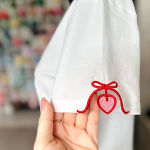 May include: White t-shirt with red embroidered trim and a pink heart on the sleeve.