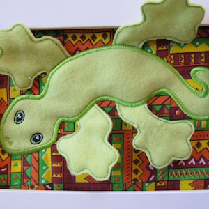 Puede incluir: Un lagarto de fieltro verde claro enmarcado con detalles bordados. El lagarto está sobre un fondo de tela estampada colorida en un marco blanco. El lagarto tiene ojos grandes y una cola curvada.