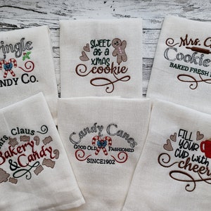 May include: Six white kitchen towels with embroidered Christmas-themed designs. The designs include "Kringle Candy Co.", "Sweet as a Xmas Cookie", "Mrs. Claus Cookie Co. Baked Fresh Daily", "Mrs. Claus Bakery & Candy", "Candy Canes Good Old Fashioned Since 1902", and "Fill Your Cup With Cheers".