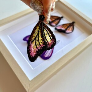 May include: A pair of butterfly-shaped earrings with black, yellow, and pink embroidery. The earrings are displayed in a white frame.