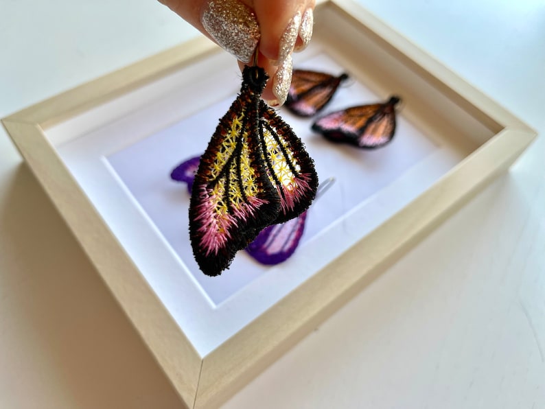 May include: A pair of butterfly-shaped earrings with black, yellow, and pink embroidery. The earrings are displayed in a white frame.