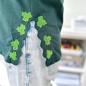 Puede incluir: Primer plano de una parte superior verde oscuro con un diseño decorativo bordado de vid y hojas en verde brillante. La parte superior se combina con unos vaqueros vaqueros de lavado claro. El bordado sigue un borde curvo.