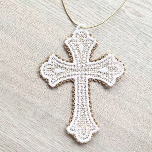 May include: White lace cross with a gold cord, hanging against a light wood background.
