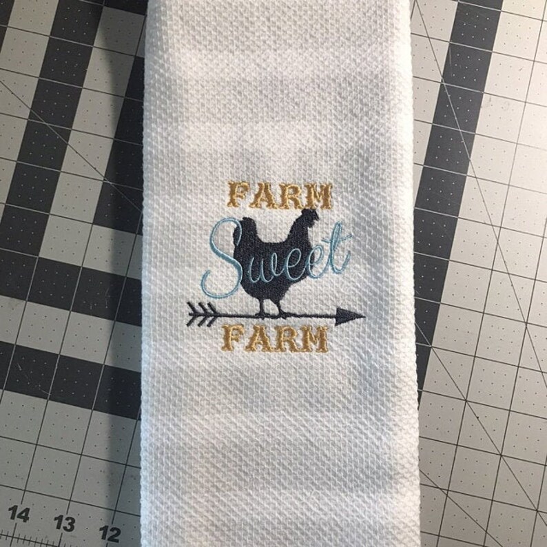 May include: White kitchen towel with embroidered text "Farm Sweet Farm" and a black silhouette of a chicken standing on an arrow.