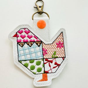 May include: A whimsical chicken-shaped keychain with a patchwork design. The bird features various patterns, including hearts, floral prints, and polka dots, in shades of pink, blue, green, and red. It has a white background, a bronze-colored clasp, and an orange button.