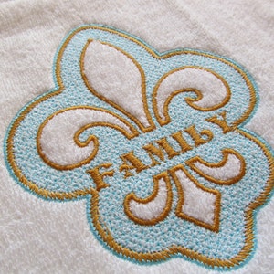 May include: White embroidered towel with a blue and gold fleur-de-lis design. The word "FAMILY" is embroidered in gold inside the fleur-de-lis.
