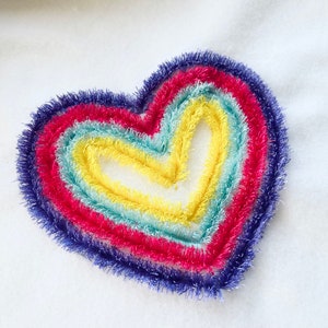 May include: A rainbow heart embroidered on a white fabric. The heart is made of five layers of yarn in different colors: purple, red, blue, green, and yellow.