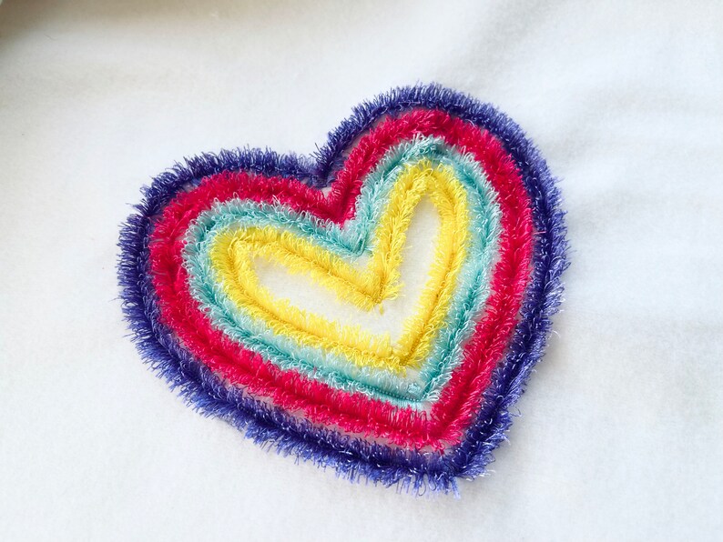 May include: A rainbow heart embroidered on a white fabric. The heart is made of five layers of yarn in different colors: purple, red, blue, green, and yellow.