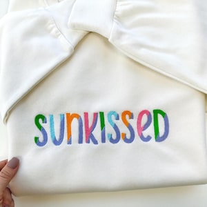 May include: White sweatshirt with the word "sunkissed" embroidered in colourful letters.