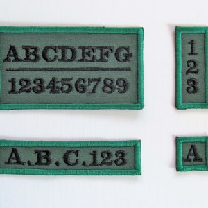 May include: Four green fabric patches with black embroidered letters and numbers. The first patch reads "ABCDEFG" and "123456789". The second patch reads "128". The third patch reads "A.B.C. 123". The fourth patch reads "A".