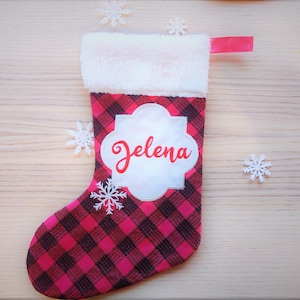Peut inclure: Chaussette de Noël en tissu écossais rouge et noir avec un revers en feutre blanc et une étiquette en feutre blanc avec le nom "Jelena" en cursive rouge.