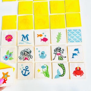 以下が含まれることがあります： 海の生き物をテーマにした刺繍生地の正方形のコレクション。デザインには、貝殻、人魚、魚、錨が含まれます。黄色の正方形も存在します。手はヒトデのデザインの正方形を持っています。