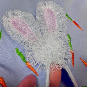 May include: Embroidered bunny rabbit design on a light blue fabric background. The bunny has white fur with pink ears and a white fluffy tail. The background is decorated with embroidered orange carrots.