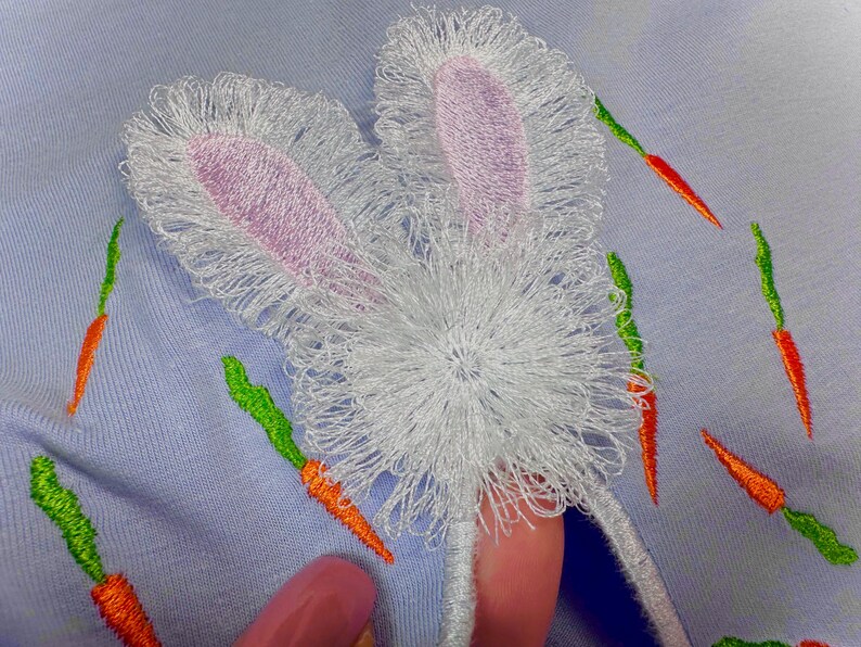 May include: Embroidered bunny rabbit design on a light blue fabric background. The bunny has white fur with pink ears and a white fluffy tail. The background is decorated with embroidered orange carrots.