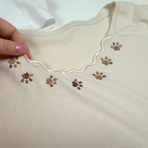 Peut inclure: Un t-shirt blanc avec un col festonné et une broderie de pattes de chat brunes.