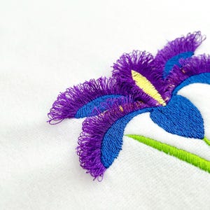 May include: Close-up of an embroidered iris flower. The flower features vibrant purple, blue, yellow, and green thread on a white background. The petals have a fringed texture, and the design is detailed.