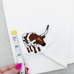May include: White fabric with an embroidered brown and white longhorn steer design. A measuring tape shows a measurement of approximately 4 inches. The fabric is folded, and the background is a light gray.