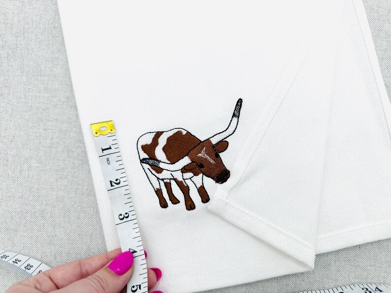 May include: White fabric with an embroidered brown and white longhorn steer design. A measuring tape shows a measurement of approximately 4 inches. The fabric is folded, and the background is a light gray.