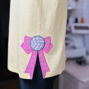 Puede incluir: Una camiseta amarilla con un aplique de lazo de voleibol rosa y gris en la manga.