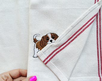 May include: A cream-colored kitchen towel featuring a folded corner and vertical red stripes. An embroidered brown and white Cavalier King Charles Spaniel puppy is visible on the fabric, peeking out from beneath the folded section of the towel.